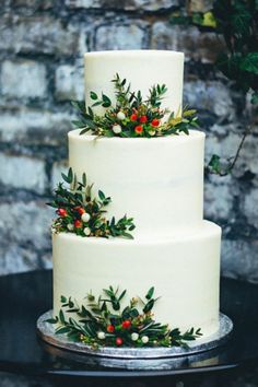 a three tiered white cake with holly and berries on the top is sitting on a black table