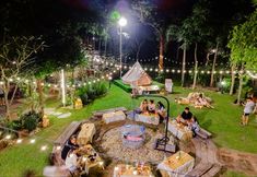 people are gathered around a fire pit in the middle of a grassy area with string lights strung across it