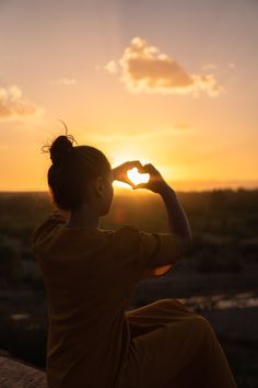 a woman sitting in front of the sun making a heart shape with her hands at sunset