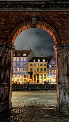 an archway leading into a city at night with buildings lit up in the background and dark clouds overhead