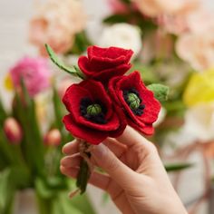 a hand holding a red felt flower in front of some pink and white flowers with green stems