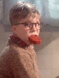 a young boy wearing glasses and a sweater with a large piece of food in his mouth