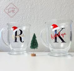 two glass mugs with christmas designs on them sitting next to a small pine tree