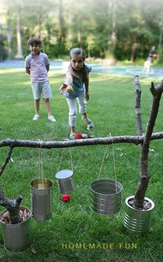 two children playing in the yard with tin cans hanging from a twig frame and some trees