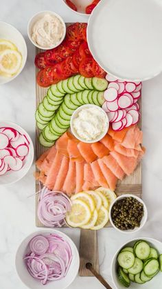 sliced up fish and vegetables on a cutting board with bowls of dips, lemons, cucumbers, tomatoes, onions, celery