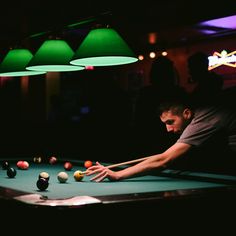 a man playing pool in a dark room with green lights hanging from the ceiling and two lamps on either side of him