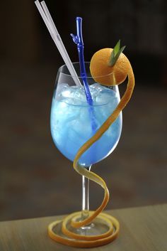 a blue cocktail with orange peels and two straws in it on a wooden table