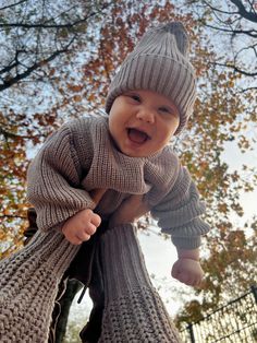 a baby in a knitted sweater and hat smiles while hanging from a tree branch