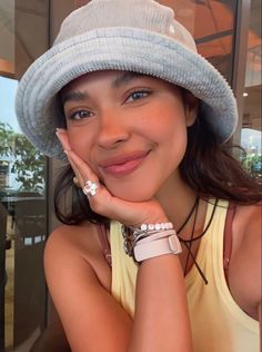 a woman wearing a white hat and bracelets smiles at the camera with her hand on her chin