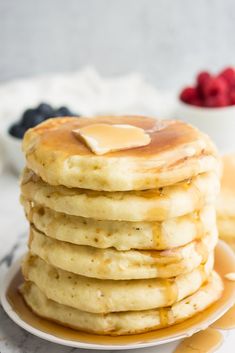 stack of pancakes with butter and syrup on white plate next to blueberries and raspberries