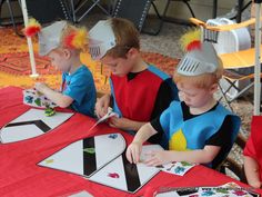 three boys sitting at a table with paper hats on their heads and one boy painting