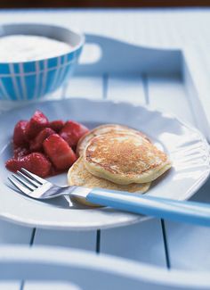 a white plate topped with pancakes and fruit next to a bowl of strawberries on a table