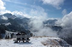 two people are sitting on a bench in the snow with mountains in the background and clouds