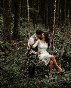 a man and woman are sitting on a swing in the woods, holding each other