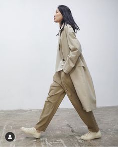 a woman walking across a cement floor wearing a trench coat and white tennis shoes in front of a white wall