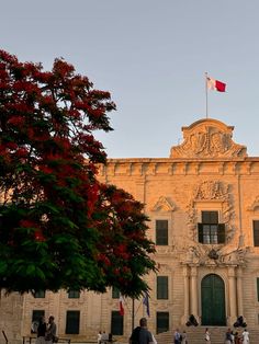 people are walking in front of an old building with a flag on the roof and trees