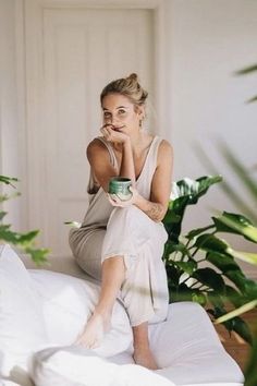 a woman sitting on top of a bed next to a potted plant and holding a cup