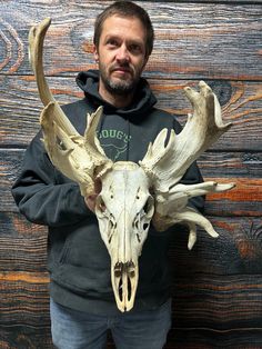 a man standing in front of a wooden wall holding a deer skull with large antlers on it