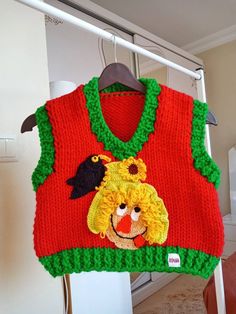 a red sweater with green trim and a bird on it is hanging from a clothes rack