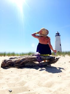 a woman sitting on top of a log in the sand near a light house and water bottle