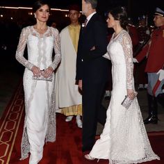 two women in white dresses standing next to each other on a red carpet with men in uniform