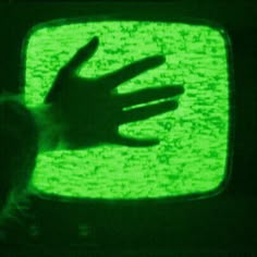 a hand on top of a television screen with green light in the dark, while someone is reaching out their arm to touch it