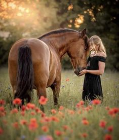 a woman standing next to a brown horse on top of a lush green field filled with red flowers