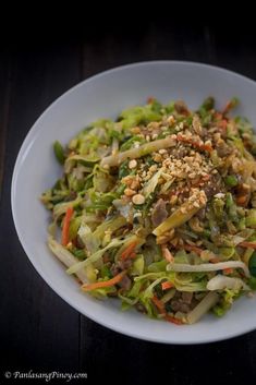 a white bowl filled with vegetables and nuts on top of a wooden table next to a green pepper