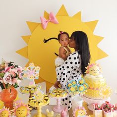 a woman holding a baby in front of a table with cakes and cupcakes