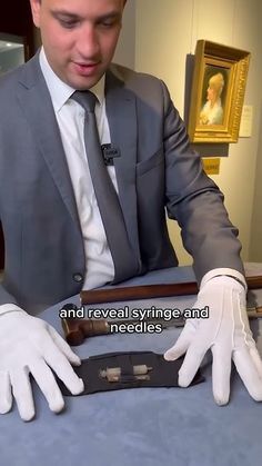 a man in a suit and white gloves is looking at something on a table with a painting behind him