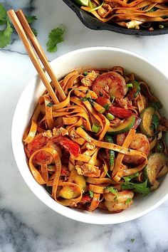 two bowls filled with noodles and vegetables on top of a marble counter next to a skillet