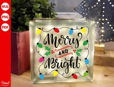 a glass block that says merry and bright with christmas lights on the front, sitting next to a potted plant