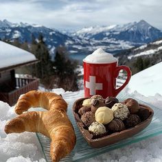 a pastry and cup of hot chocolate on a tray in the snow with mountains in the background