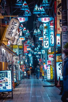 people are walking down an alley way at night with neon signs on the buildings above them