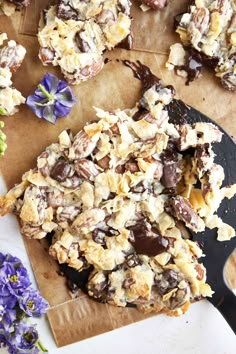 a cookie with chocolate chips and nuts on a piece of parchment paper next to purple flowers