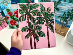 a person holding up a pink painting with palm trees on it in front of other paintings