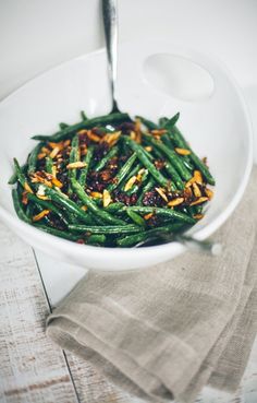 a white bowl filled with green beans on top of a wooden table next to a spoon