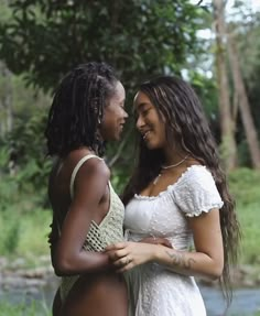 two beautiful women standing next to each other in front of a river with trees behind them