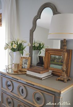 a dresser with flowers and pictures on it in front of a mirror, lamp and window