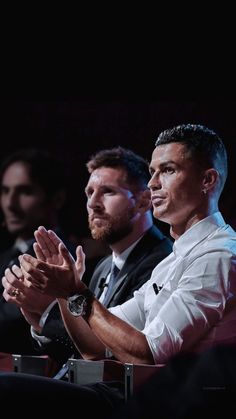 two men sitting in front of a crowd clapping hands while one man is wearing a white shirt and tie