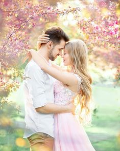 a man and woman standing under a tree with pink flowers in the foreground, looking into each other's eyes