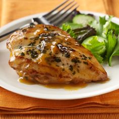 a white plate topped with chicken next to a green salad and fork on top of a wooden table