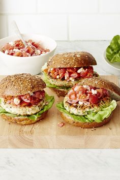 three sandwiches with lettuce, tomato and cheese sitting on a cutting board next to a bowl of salad