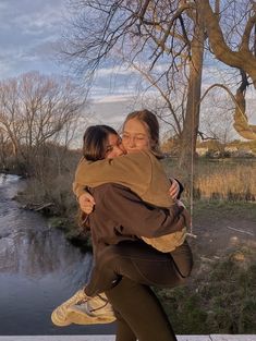 two women hugging each other in front of a river and trees with no leaves on them