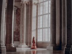 a woman in a red dress is walking through an empty room with columns and large windows