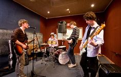 three young men are playing instruments in a recording studio while one man is singing into the microphone