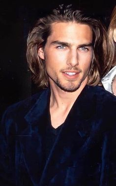 a man with long hair wearing a dark blue suit and black shirt is looking at the camera