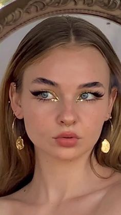 a woman with blue eyes and gold make - up looks into the camera in front of a mirror