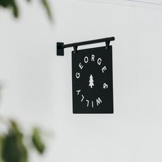 a black sign hanging from the side of a white wall next to a green leafy tree