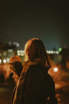 a woman standing on the street at night with her back turned to the camera and lights in the background